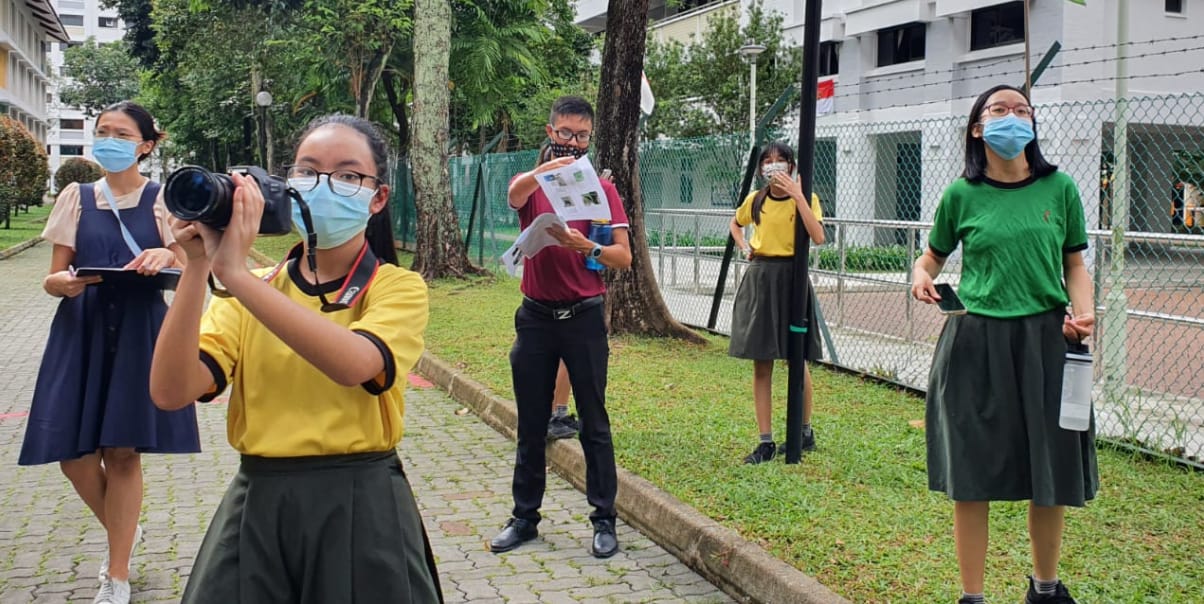 Five masked students in school uniforms stand outdoors, one holding a camera and another holding printed sheets.